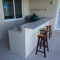 Chic outdoor bar setup with a granite countertop, stylish wooden stools, and a ceramic smoker, showcasing Palm Bay's craftsmanship in patio entertainment spaces.