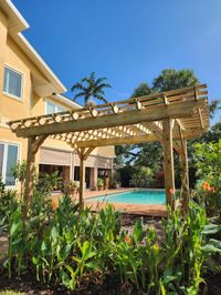 Elegant backyard wooden pergola casting gentle shadows over a lush garden with blooming flowers, adjacent to a pool in a sunny Palm Bay home.