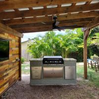 Stylish outdoor kitchen under a pergola with ceiling fan, featuring a built-in grill and stainless steel cabinets, by Palm Bay's custom exteriors service.