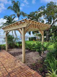 Picturesque wooden pergola with open lattice roof over a brick pathway, tropical palm trees in the background, enhancing a waterfront property in Florida.