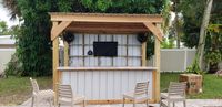 Outdoor kitchen bar with rustic wooden overhang, mounted TV, and speakers, perfect for entertaining in a Florida backyard setting.